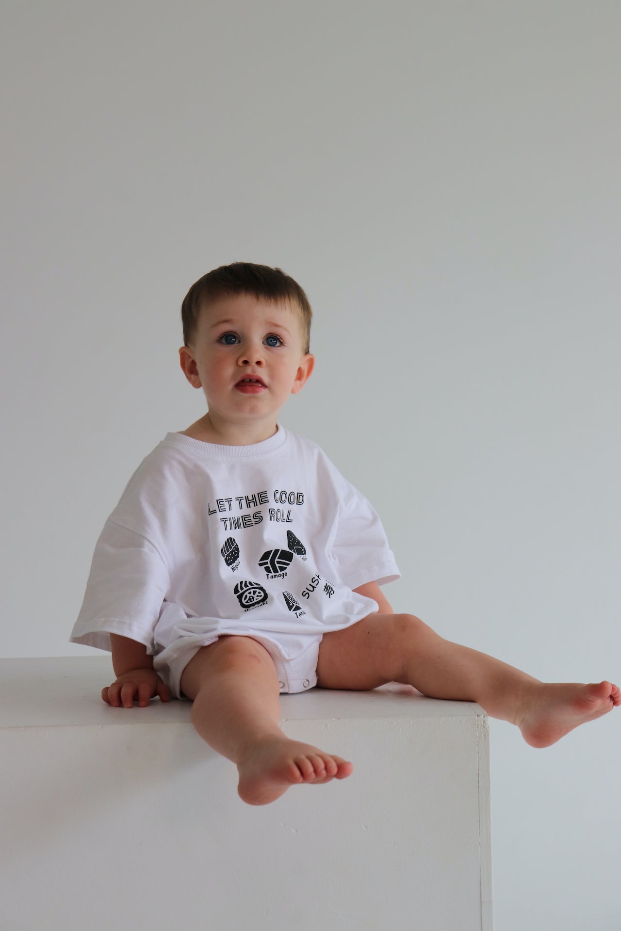 Child wearing a white oversized romper with a black sushi logo 'Let the good times roll'