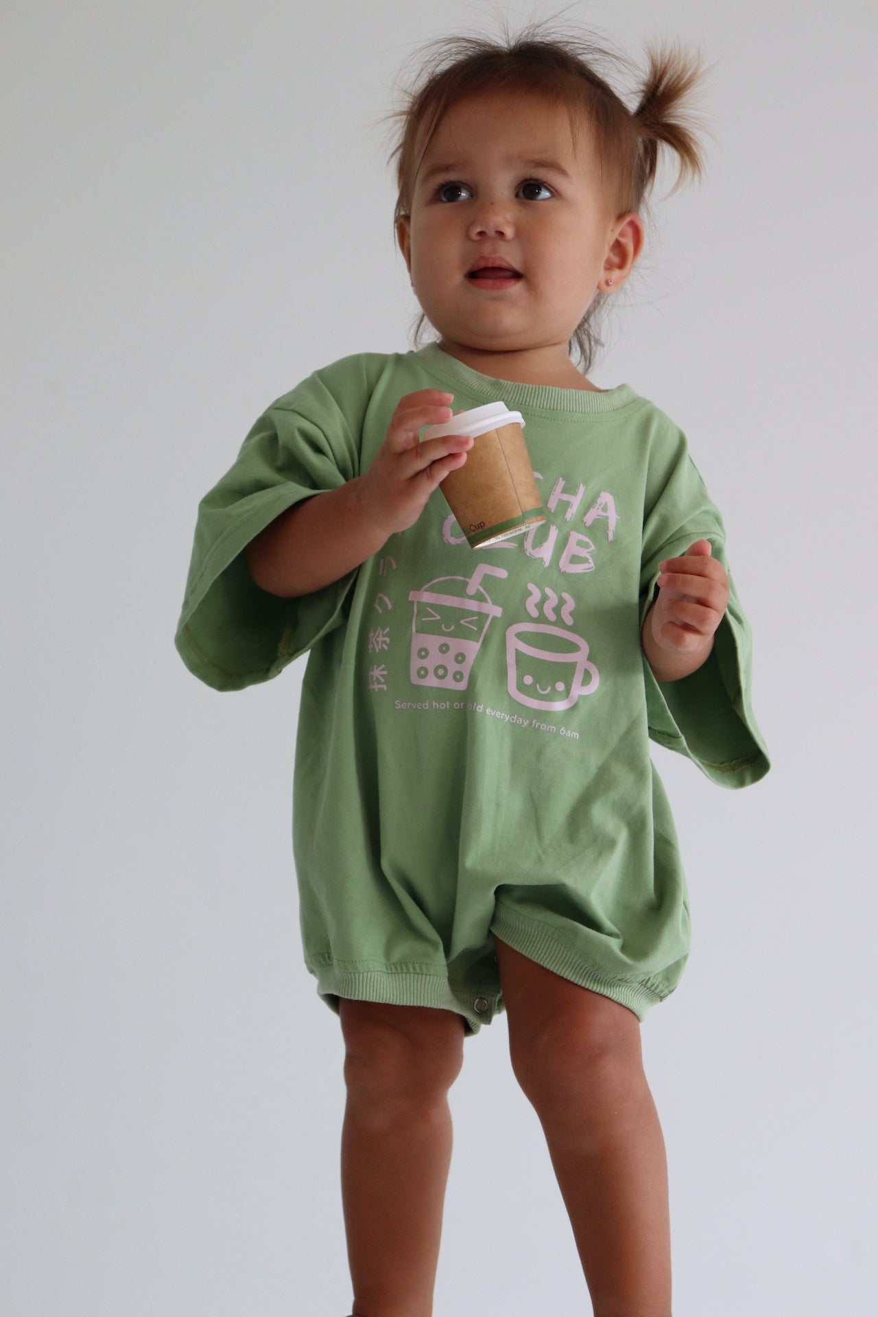 Child wearing an oversized green romper with a pink matcha club logo.