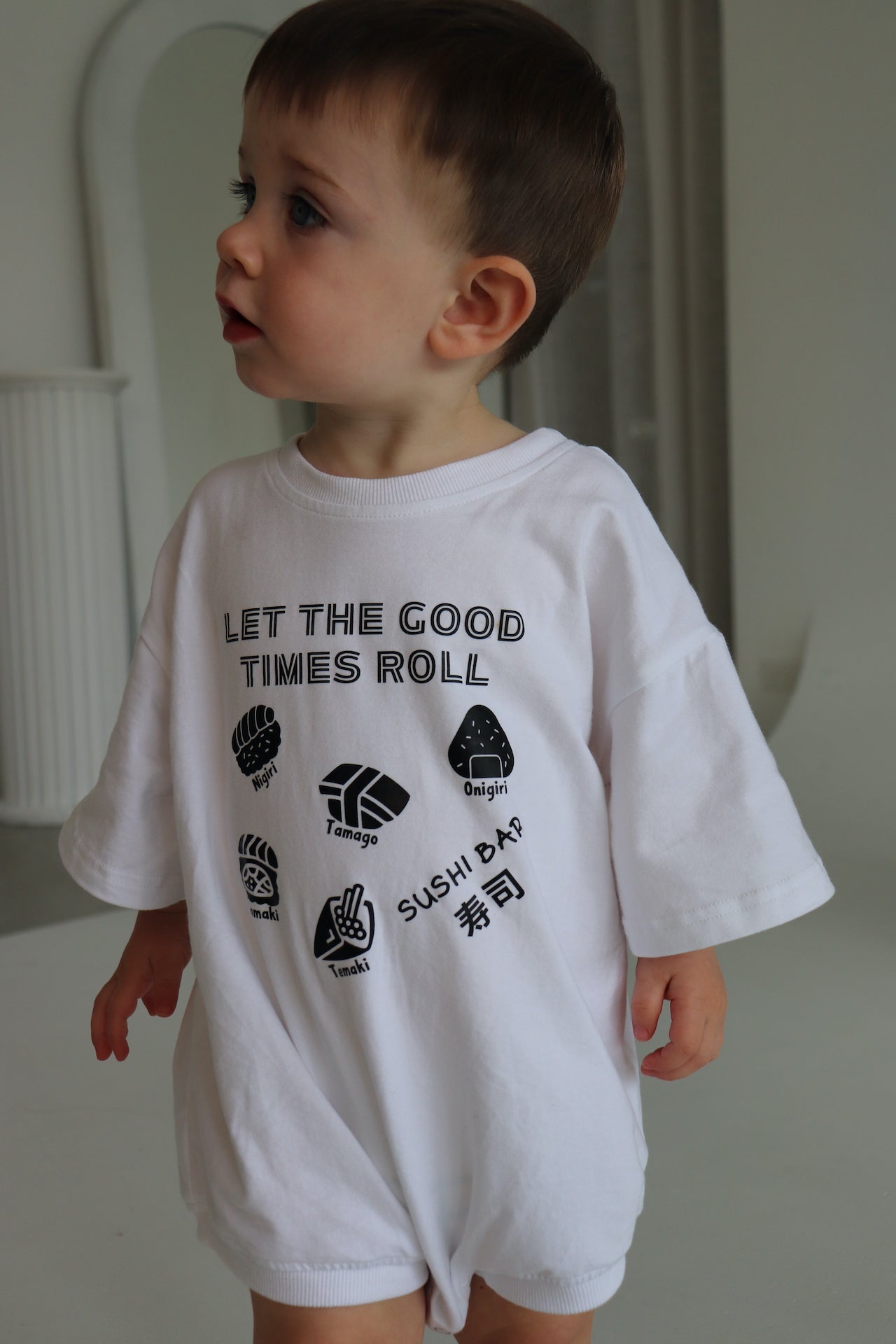 Child wearing a white oversized romper with a black sushi logo 'Let the good times roll'