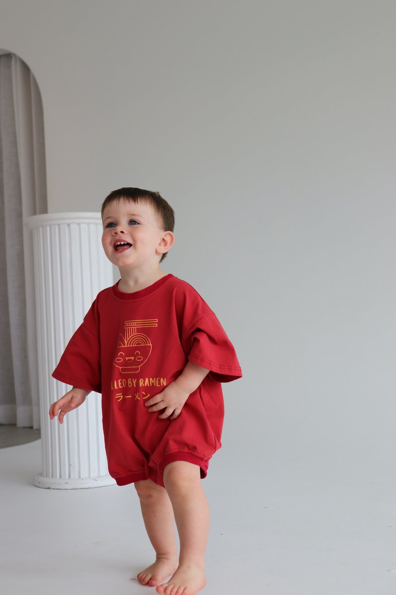 Child wearing an oversized red romper with an orange ramen bowl logo with 'Fuelled by ramen' writing