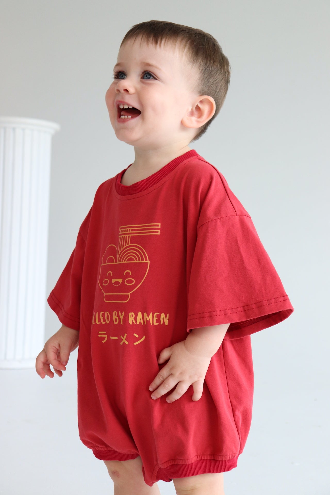 Child wearing an oversized red romper with an orange ramen bowl logo with 'Fuelled by ramen' writing.