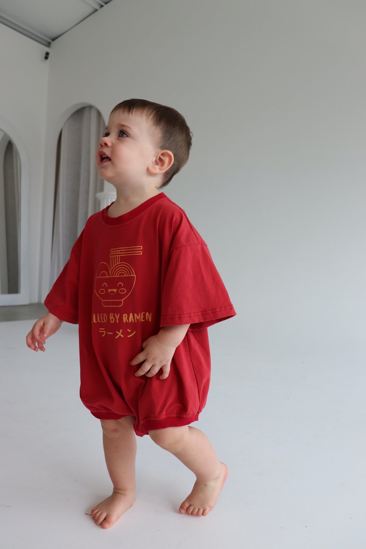 Child wearing an oversized red romper with an orange ramen bowl logo with 'Fuelled by ramen' writing