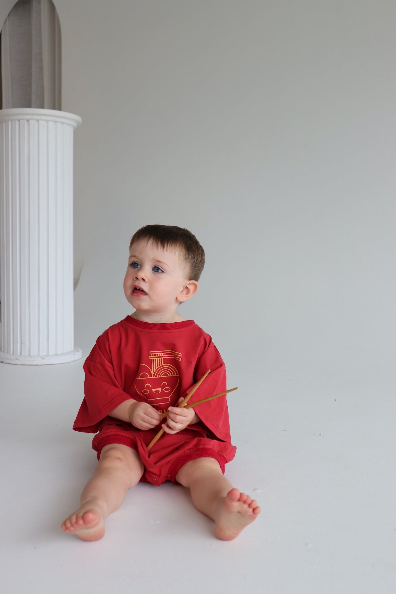 Child wearing an oversized red romper with an orange ramen bowl logo with 'Fuelled by ramen' writing