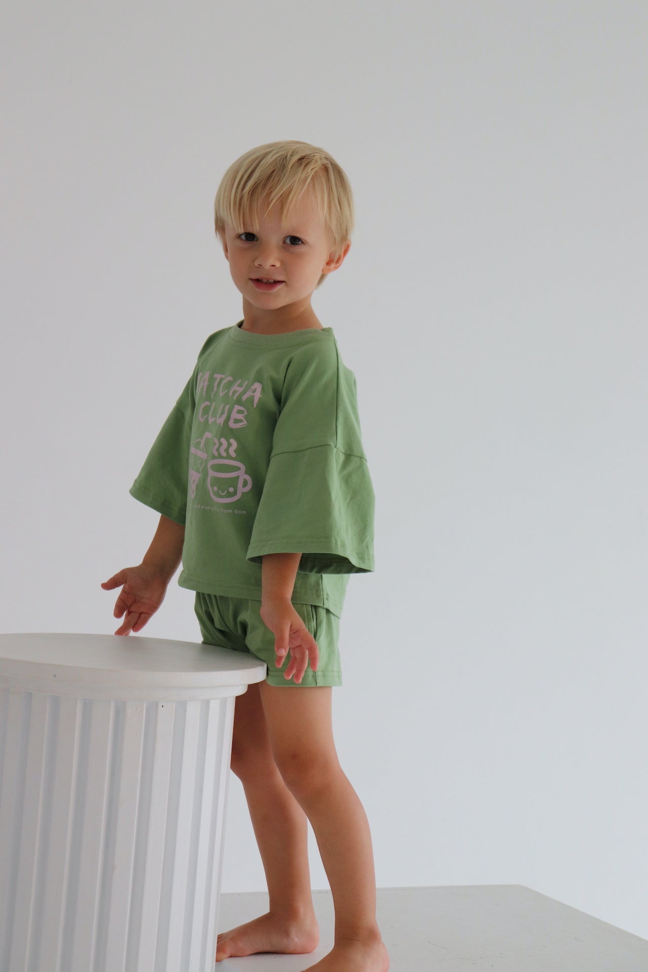Child wearing an oversized green t-shirt set with a pink matcha club logo.