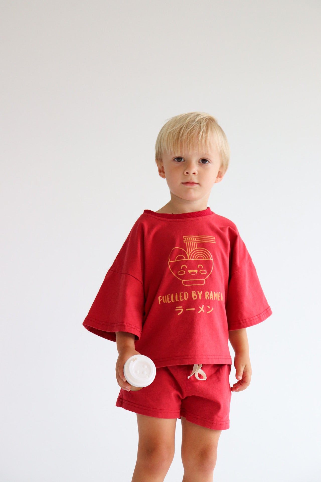 Child wearing an oversized red t shirt and short set with an orange ramen bowl logo with 'Fuelled by ramen' writing